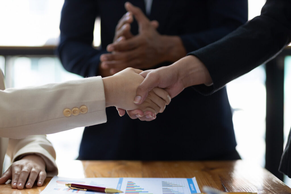 Two business professionals shaking hands after closing a deal, with one of their colleagues clapping hands in the background.