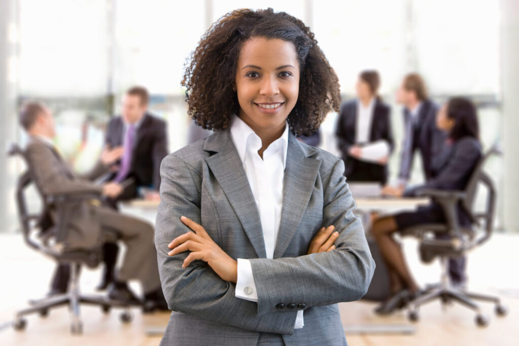 Young corporate professional standing confidently with her team discussing in the background.